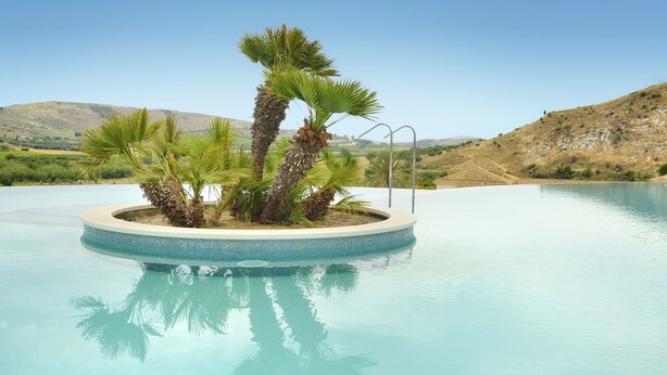 A swimming pool with a circular island featuring palm trees, surrounded by a clear blue sky and a hilly landscape. - Baglio Caruana Cantina & Relais (Siculiana)