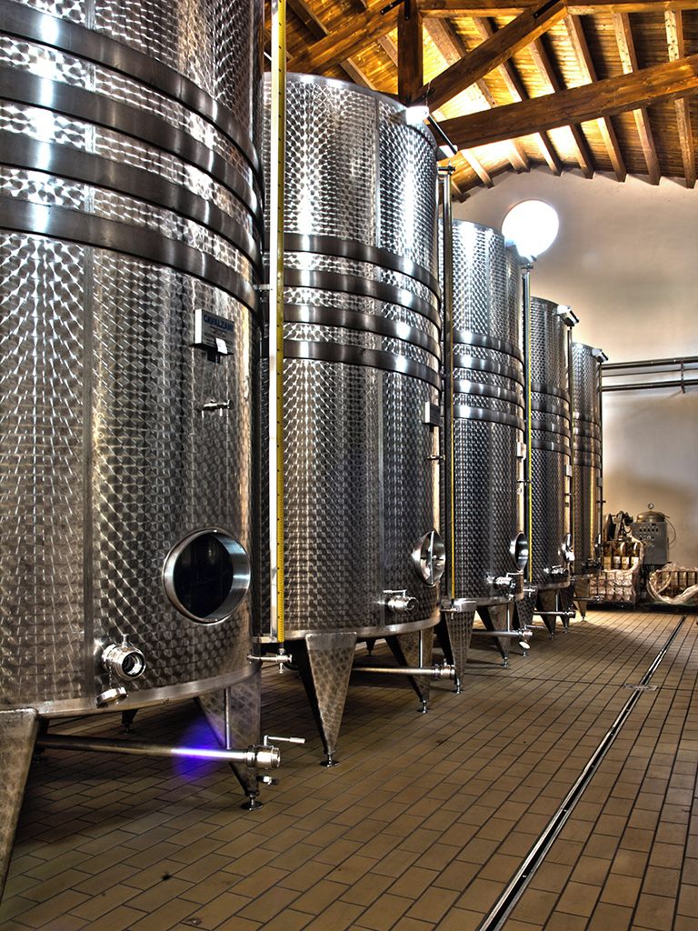 Stainless steel fermentation tanks in a warehouse setting with a wooden ceiling and brick floor.