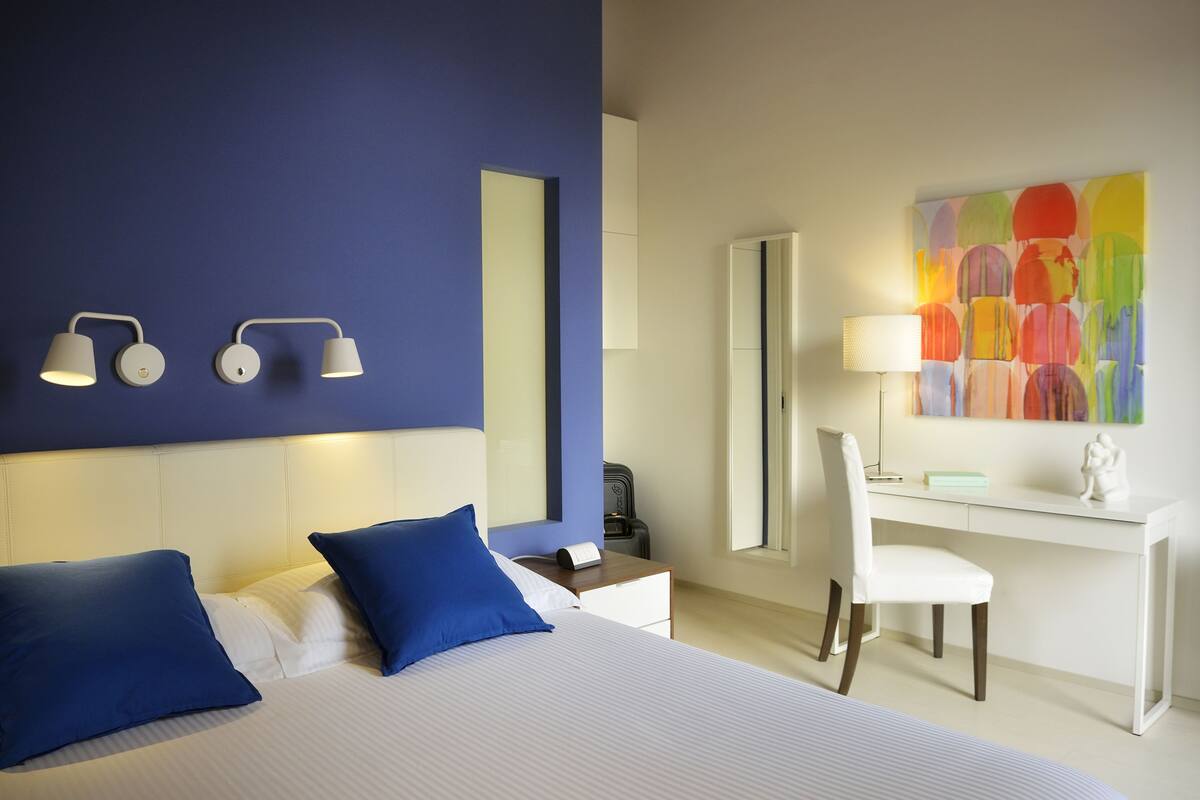 Ein Hotelzimmer mit Bett, Schreibtisch, Stuhl, Lampe und einem farbenfrohen abstrakten Gemälde.