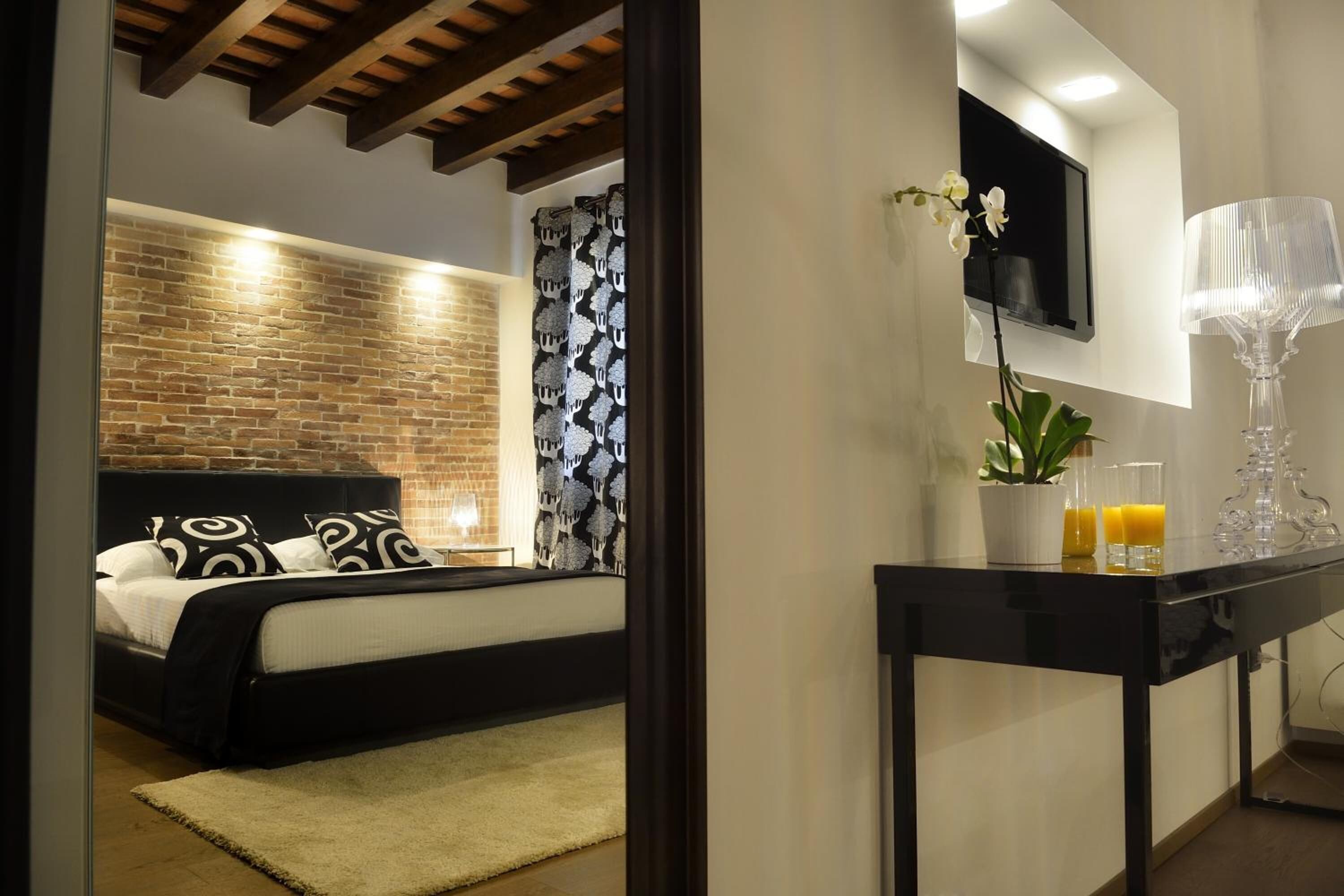 A bedroom with a brick wall, a bed with black and white bedding, a TV mounted on the wall, a small table with a plant and glasses, and a lamp.