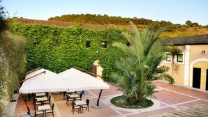An outdoor patio area with tables and umbrellas, surrounded by lush greenery and a building with a tiled roof. - Baglio Caruana Cantina & Relais (Siculiana)