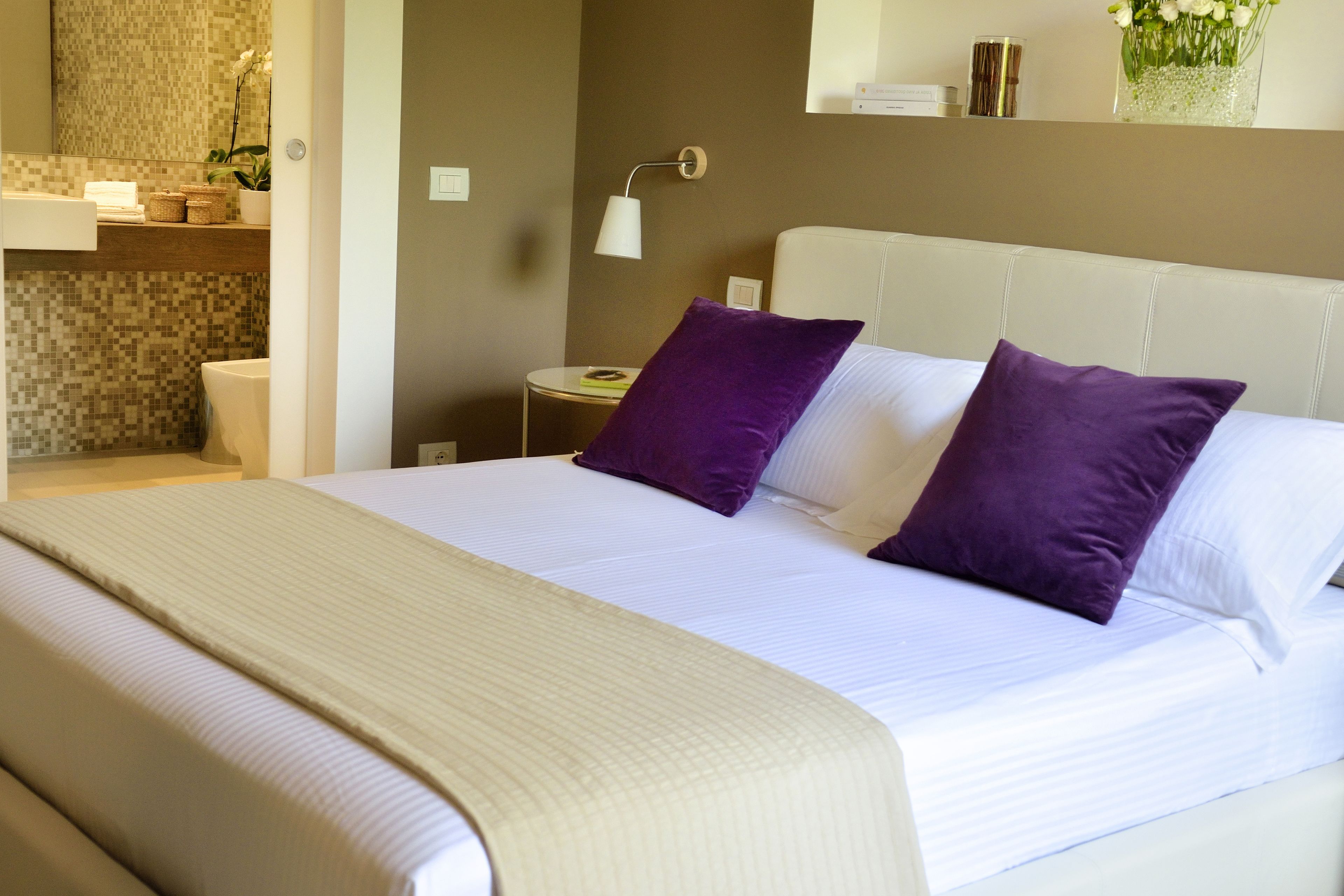 A neatly made bed with white linens and two purple pillows, a bedside table with a lamp, and a small shelf with toiletries.