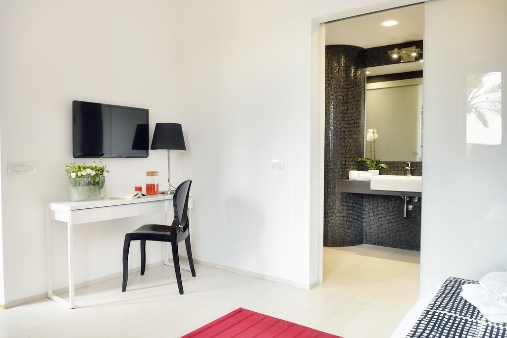 Una habitación de hotel moderna con un escritorio blanco, una silla negra, un televisor de pantalla plana, una alfombra roja y un baño con pared de azulejos negros y un espejo grande.