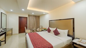 A hotel room with a large bed, a desk with a chair, a small table with a vase of flowers, and a telephone on the nightstand. - Hotel Lance International (Nagercoil)