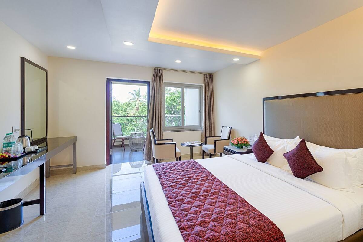 A hotel room with a large bed, a desk with a telephone, and a balcony with chairs.