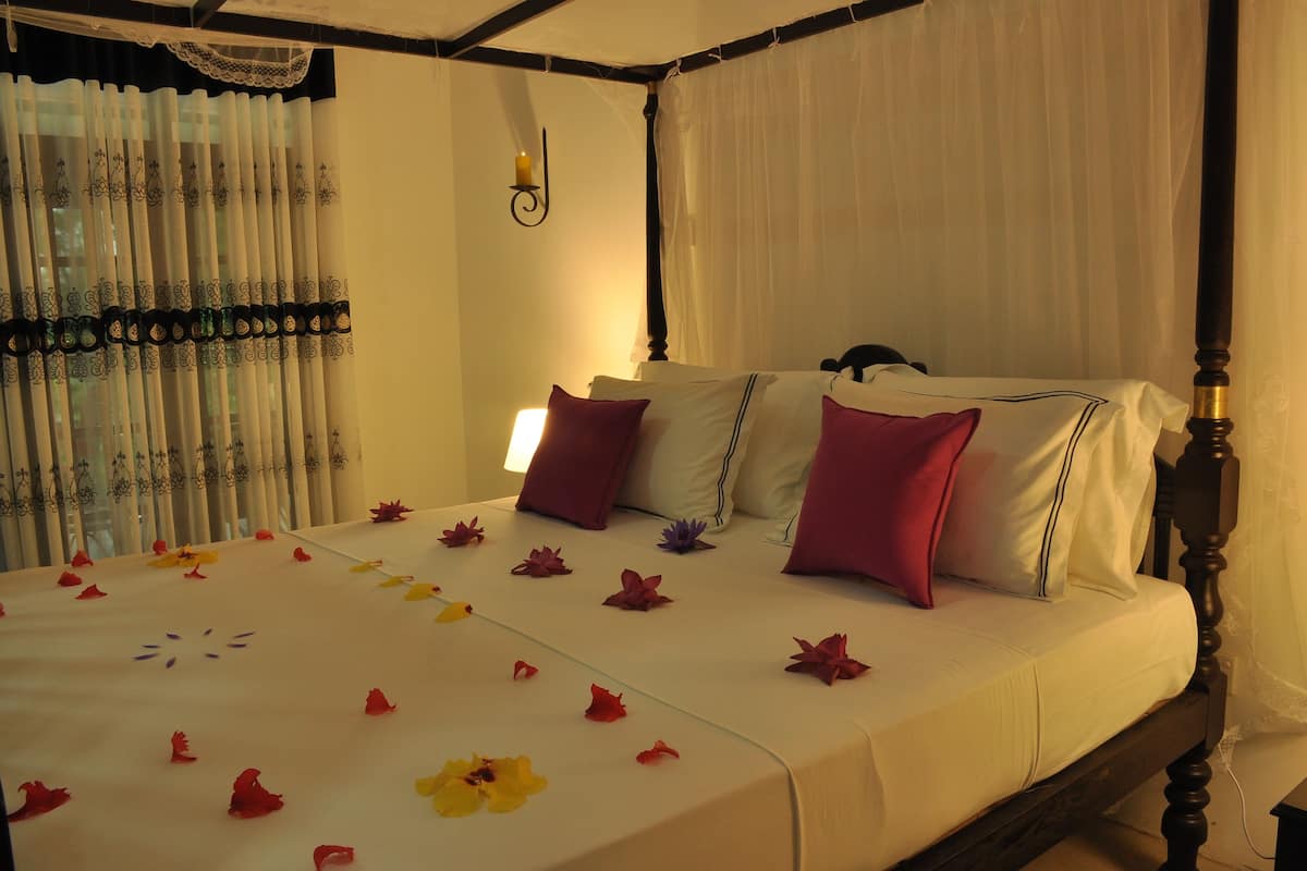 A neatly made bed with a white bedspread and purple pillows, a bedside table with a lamp, and a window with sheer curtains.