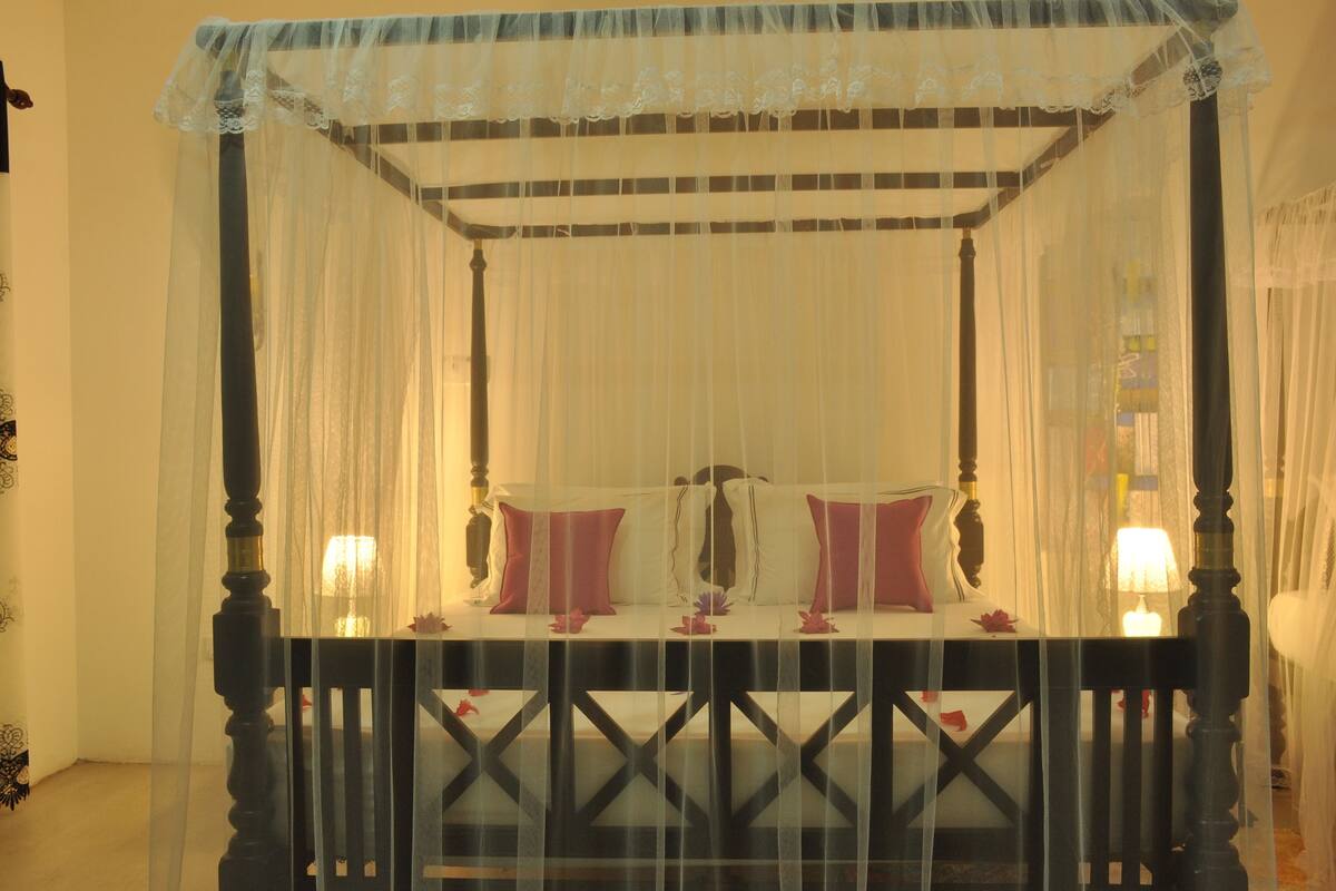 A four-poster canopy bed with a white bedspread and red pillows, a wooden headboard, and a nightstand with a lamp.
