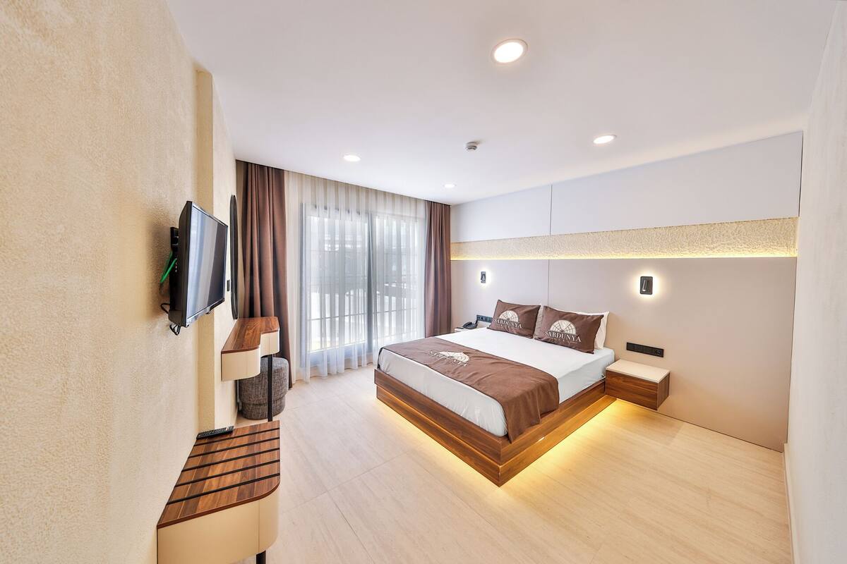 A modern hotel room with a large bed, a flat-screen TV mounted on the wall, a bedside table, and a desk with a chair.
