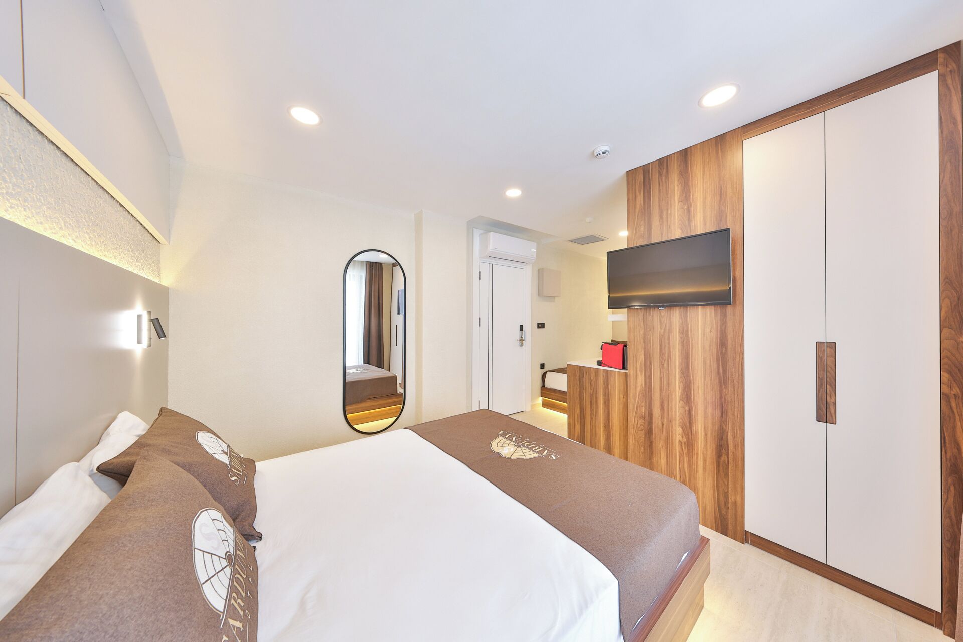 A hotel room with a bed, a mirror, a TV mounted on the wall, and a wardrobe.