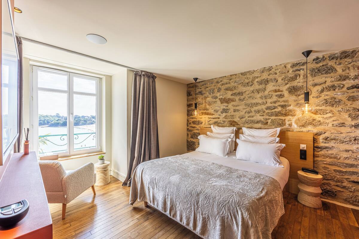 A bedroom with a large bed, a desk with a bowl, a chair, a window with curtains, and a stone wall.