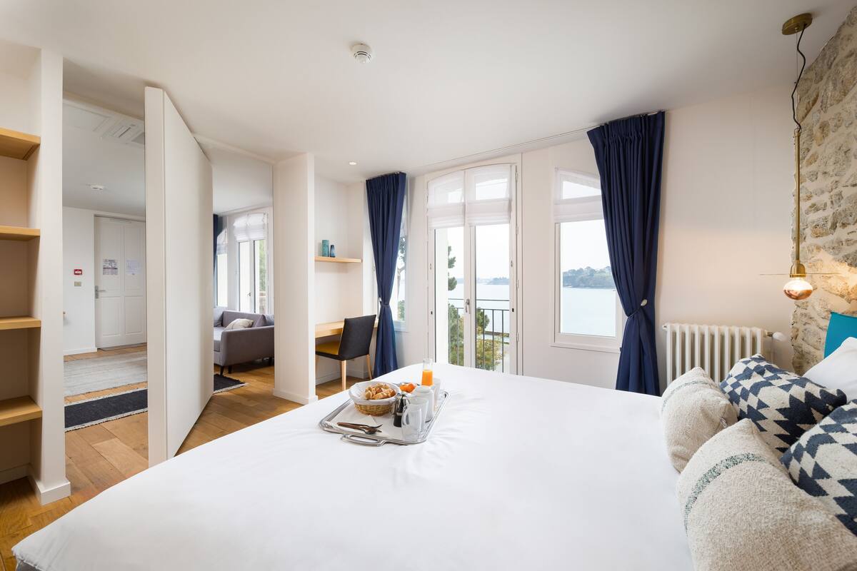 A hotel room with a large bed, breakfast tray, a view of the outdoors, a balcony, and a stone wall.