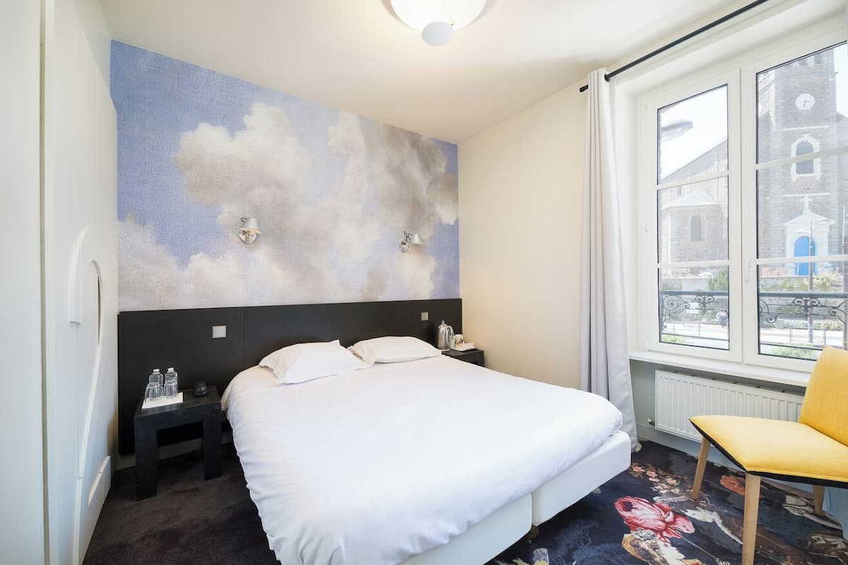 A hotel room with a bed, a nightstand, a chair, a window with a view of buildings, and a wall with a cloud mural.