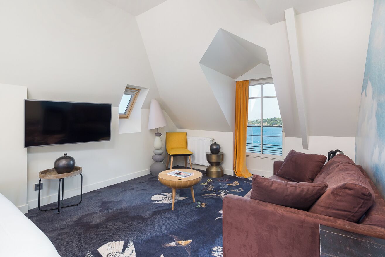 A modern hotel room with a flat-screen TV, a sofa, a small round table, and a view of the sea through large windows.