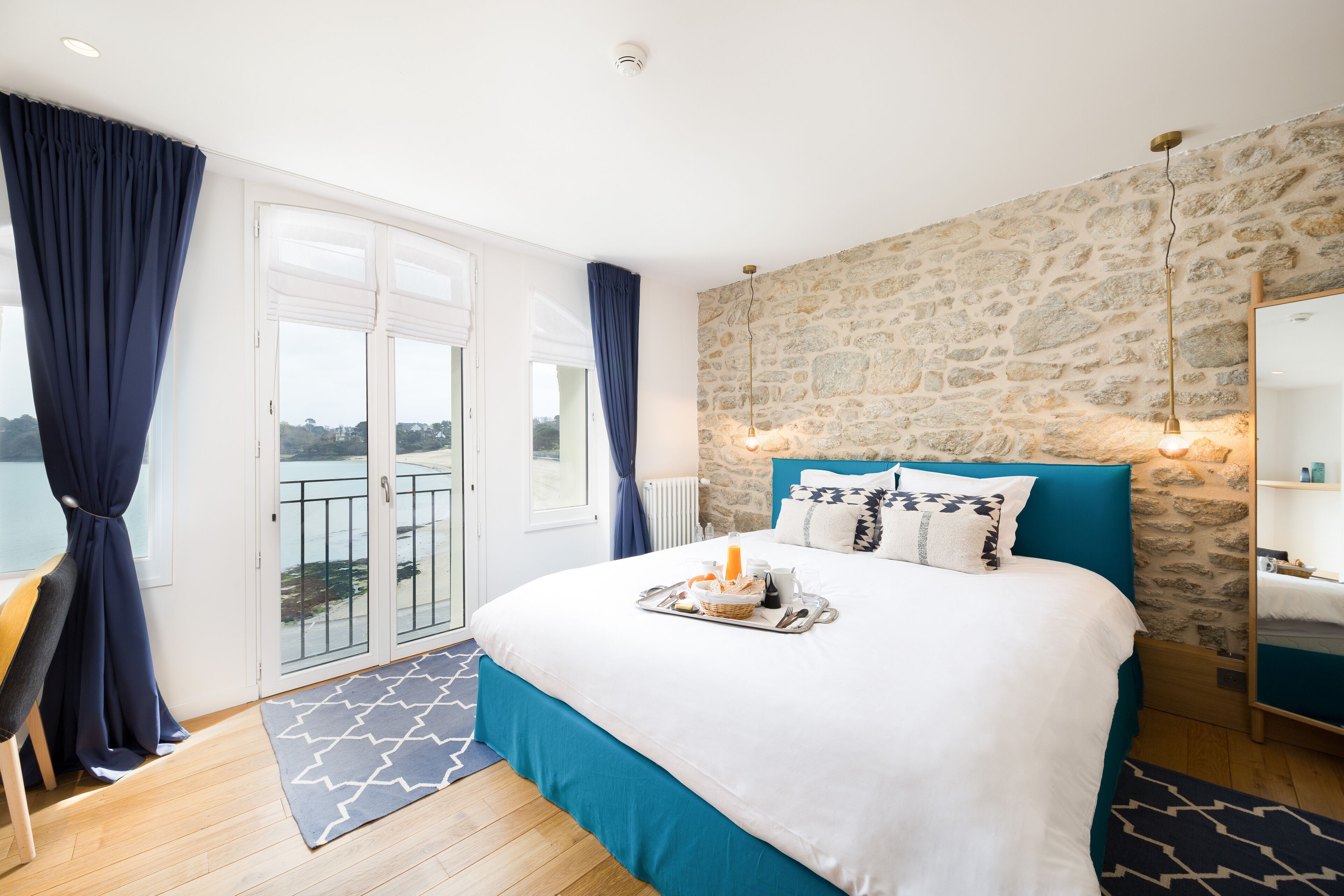 Un dormitorio con pared de piedra, una cama con cabecera azul, una bandeja con alimentos para el desayuno y vistas a través de un ventanal.