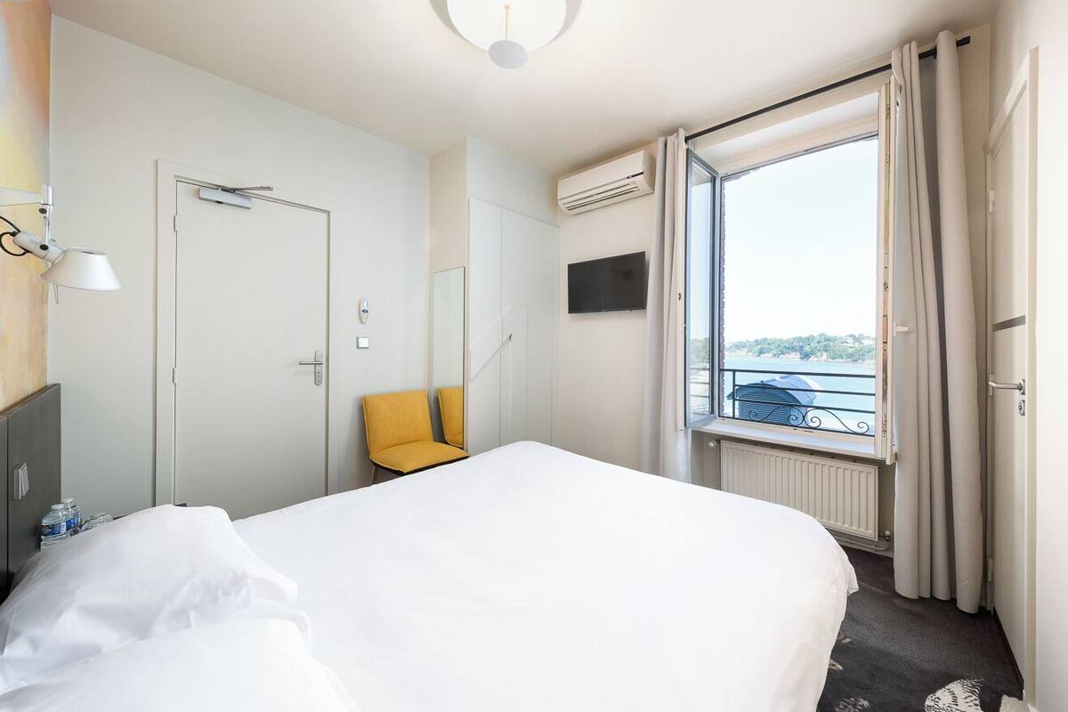 A hotel room with a bed, a chair, a desk with a lamp, an air conditioning unit, a TV, and a window with curtains.