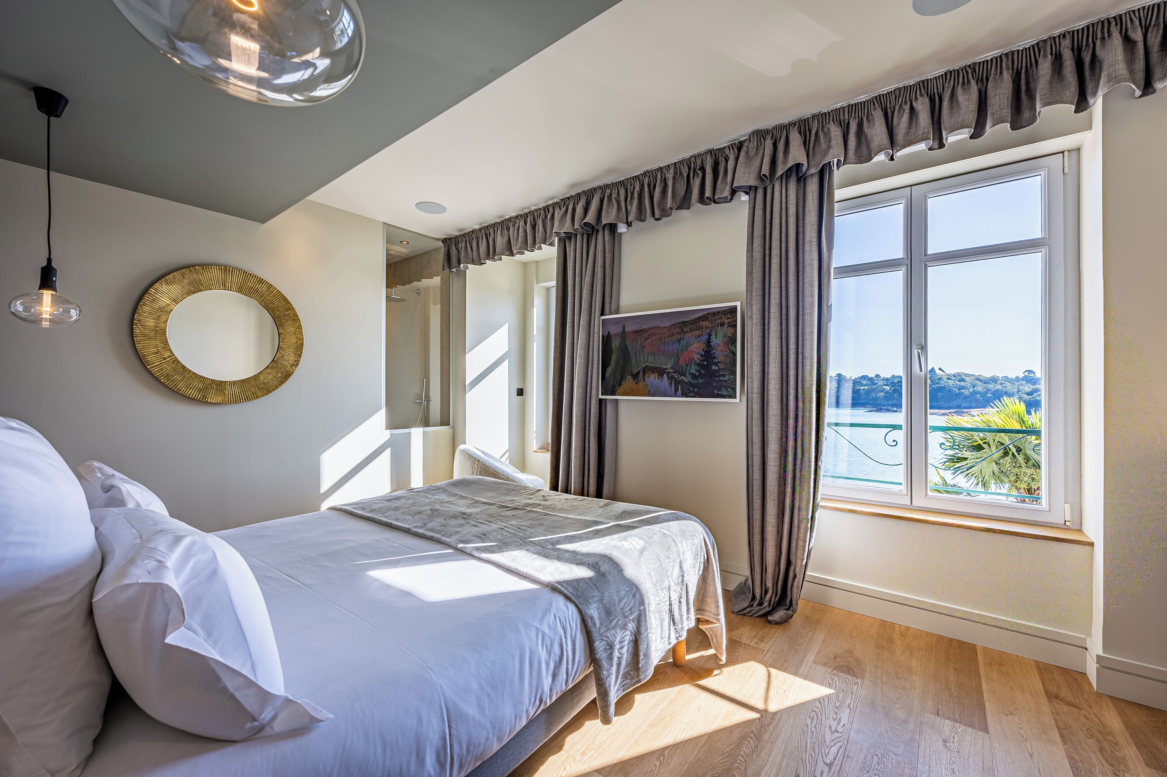 A bedroom with a large bed, a round mirror, a painting, and a view of greenery through the window.