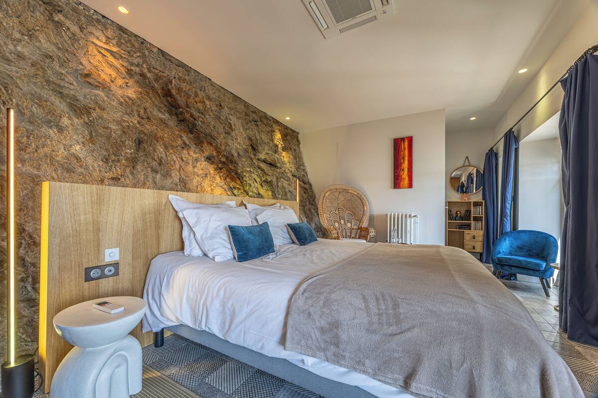 A modern bedroom with a large bed, a bedside table, a wall-mounted heater, a wicker chair, and a view of the room.