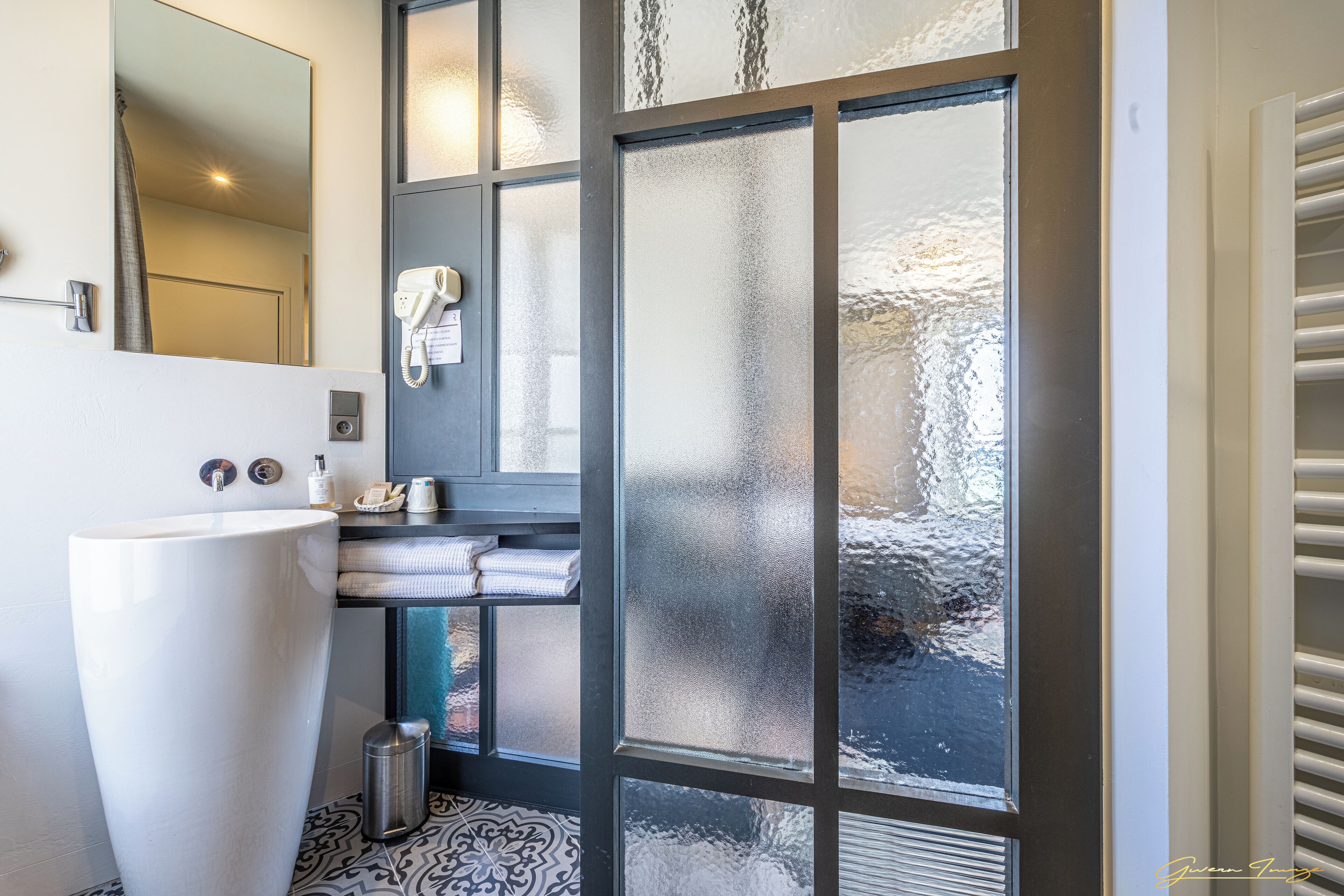 Un baño con un lavamanos grande y blanco, un espejo, un perchero para toallas y una puerta de cristal con paneles satinados.