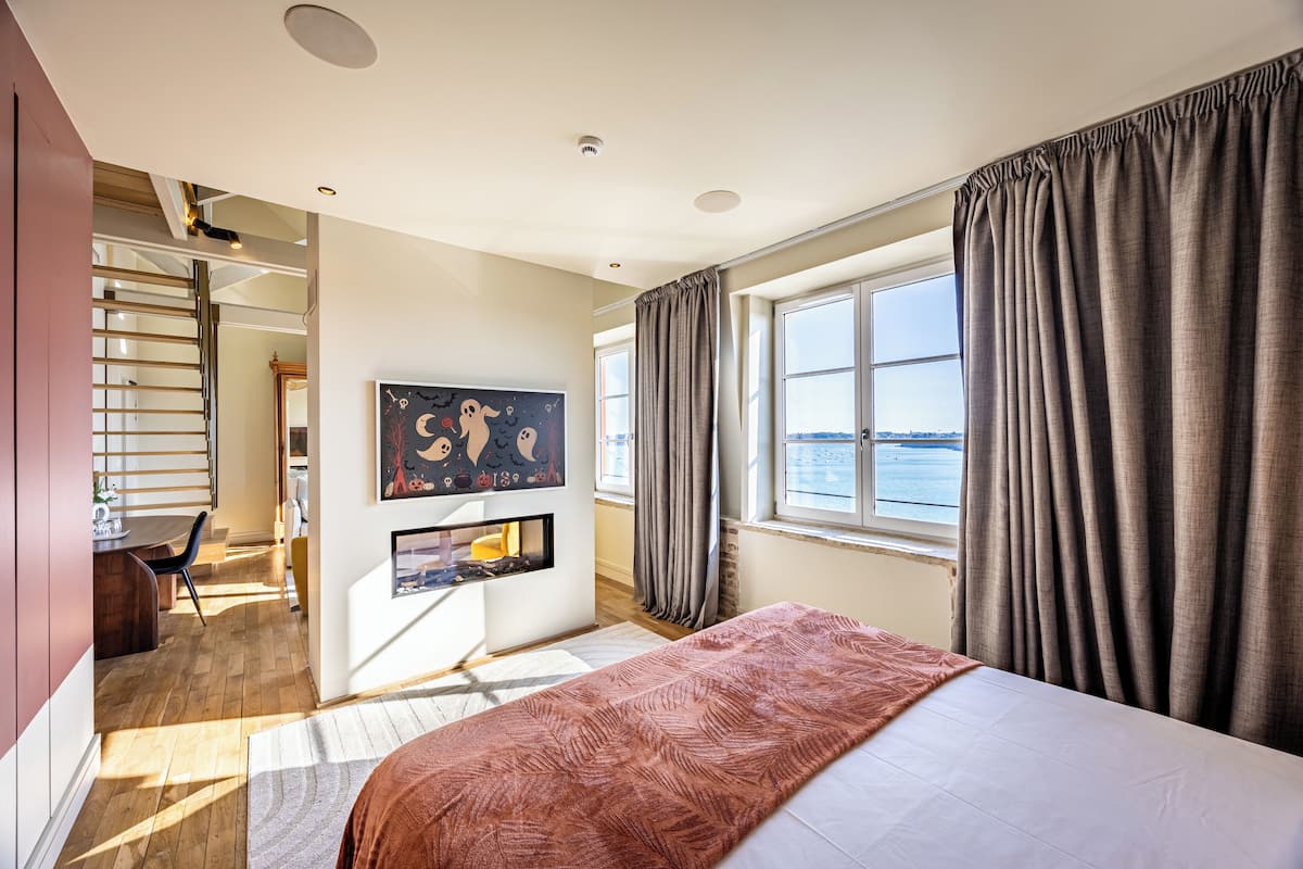 A hotel room with a bed, a desk, a chair, a painting, a window with curtains, and a view of the sea.