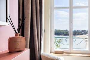 Une pièce avec vue sur une étendue d’eau, visible par une fenêtre, un vase contenant des tiges noires et une petite table sur laquelle se trouve une boîte.