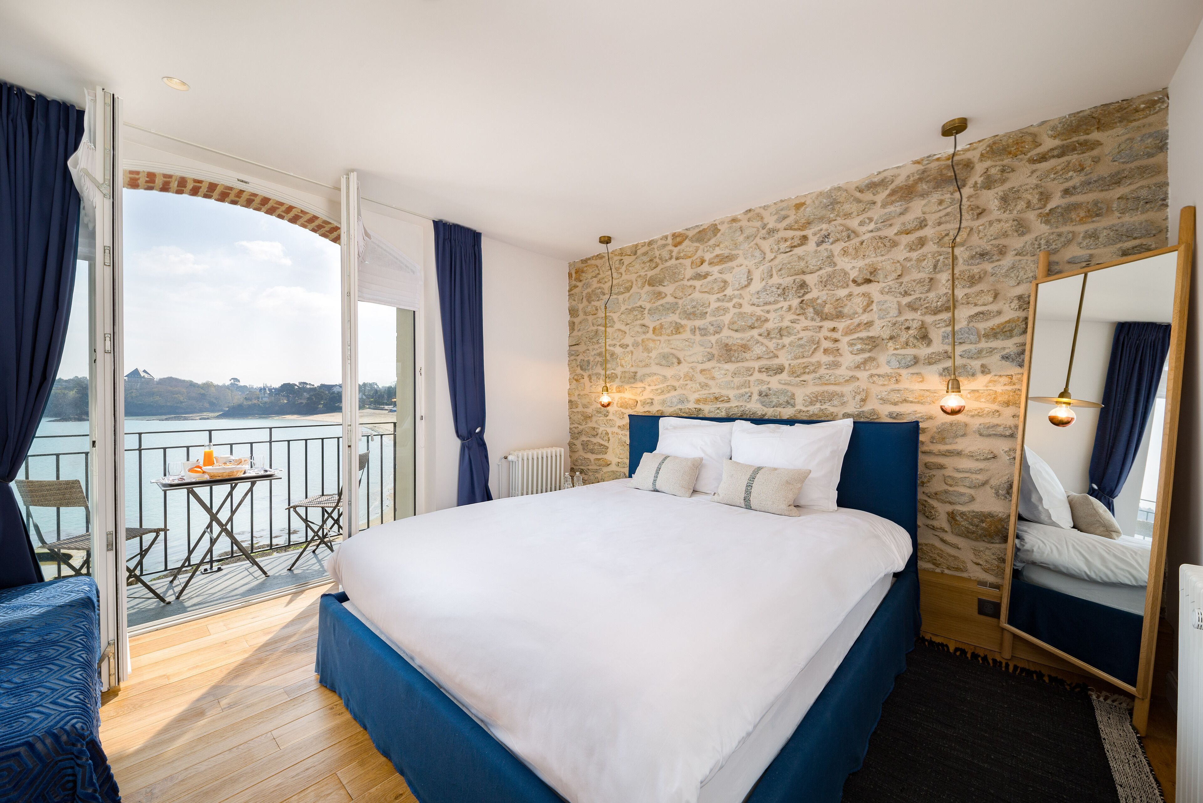 Un dormitorio con una cama grande, una pared de piedra, un balcón con mesa y sillas, y vista al mar.