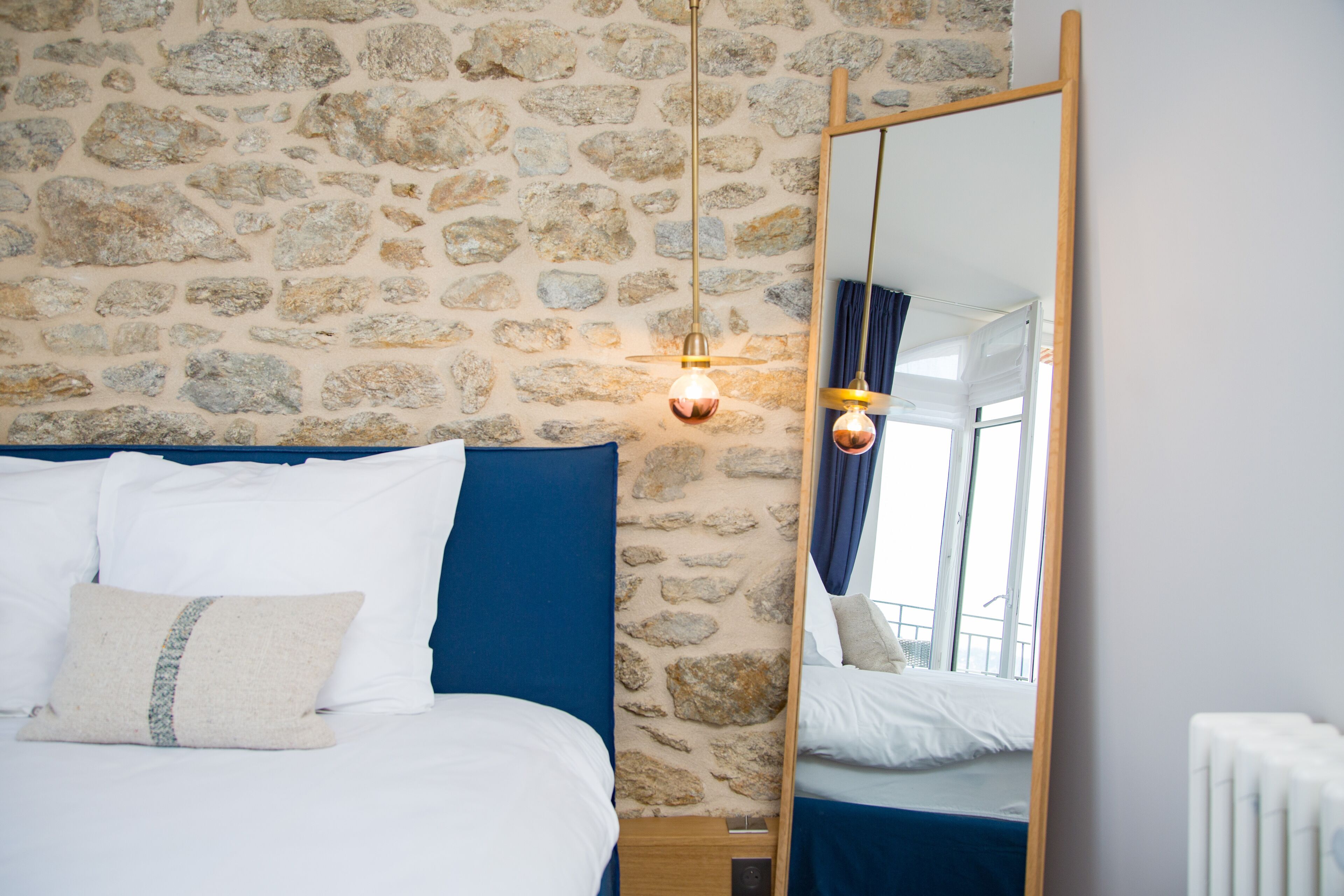 A bedroom with a stone wall, a bed with white and blue bedding, a wooden headboard, a hanging light fixture, and a mirror reflecting the room.