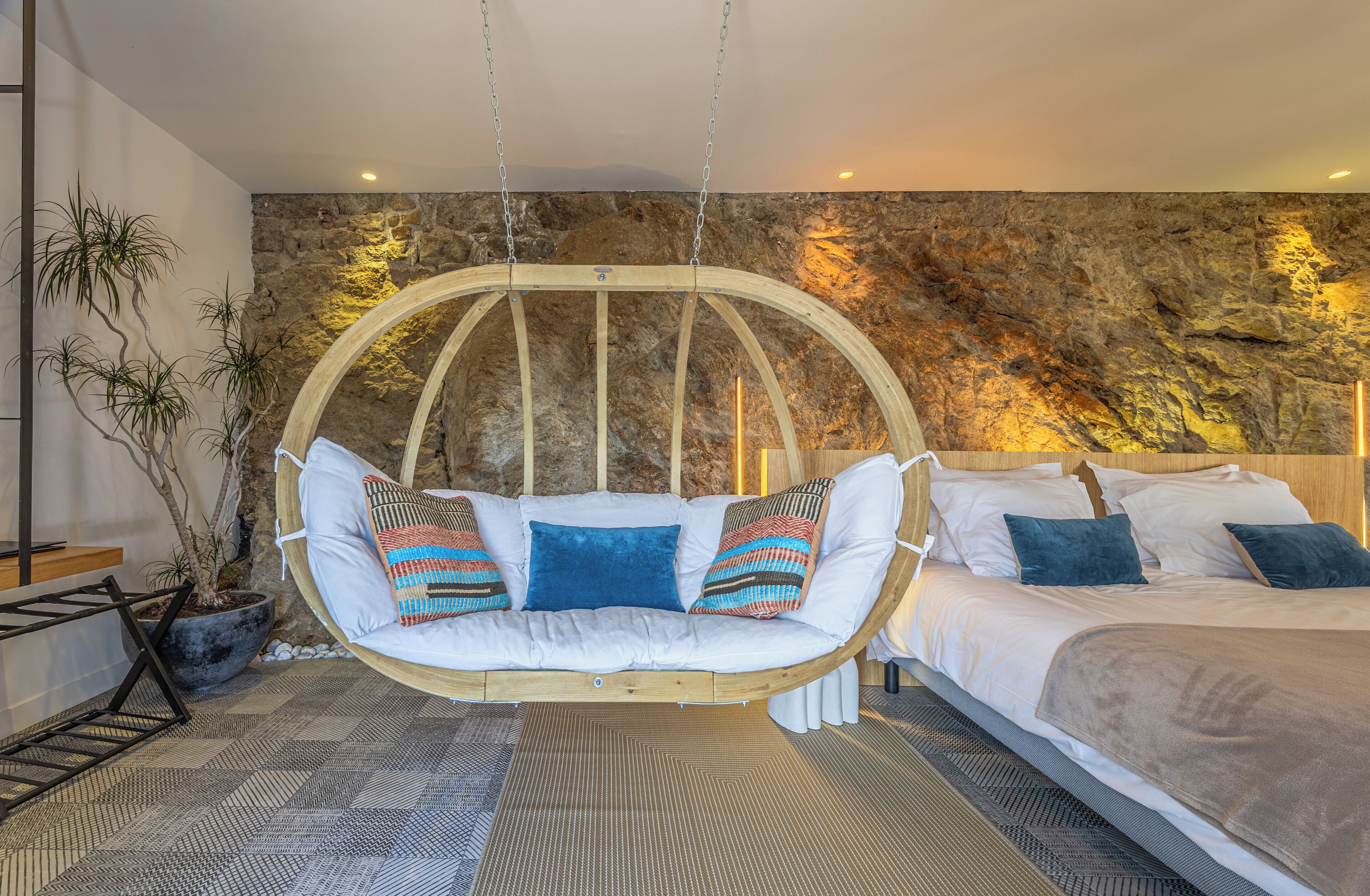 A bedroom with a hanging chair, a bed, and a stone wall.