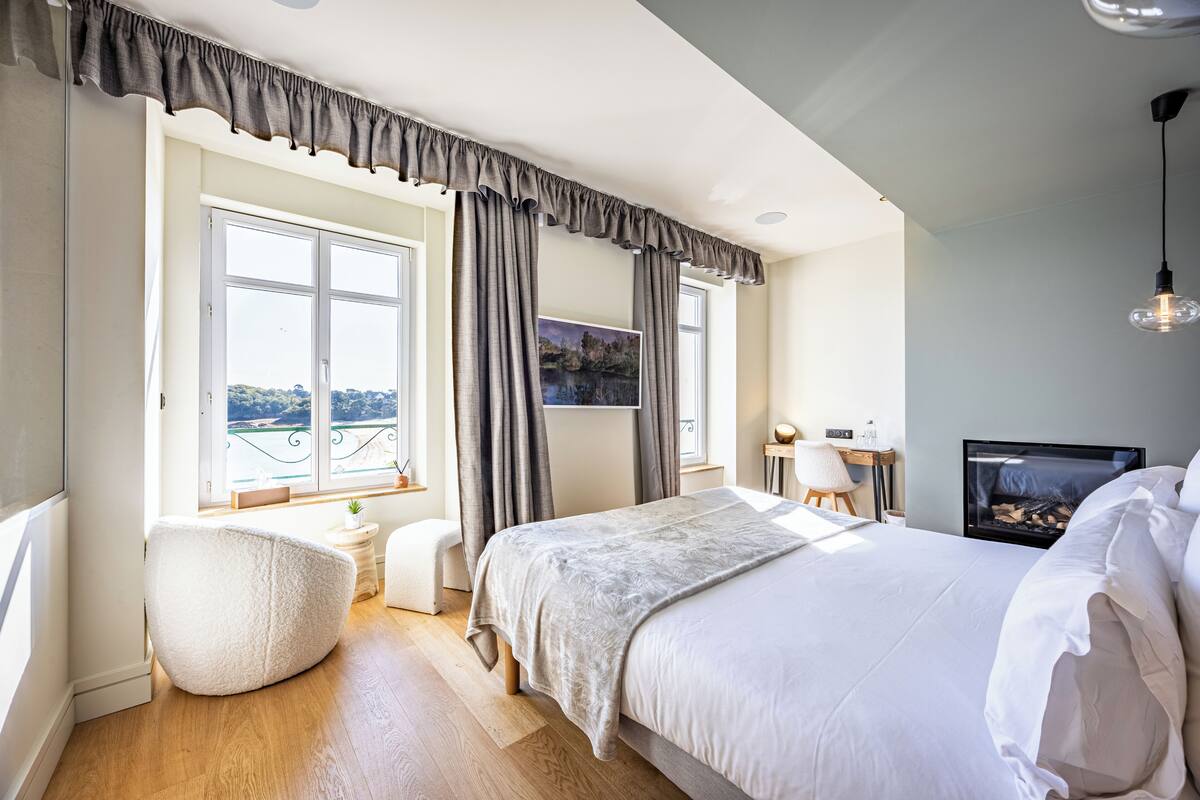 A bedroom with a large bed, a window with curtains, a small table with a lamp, and a fireplace.