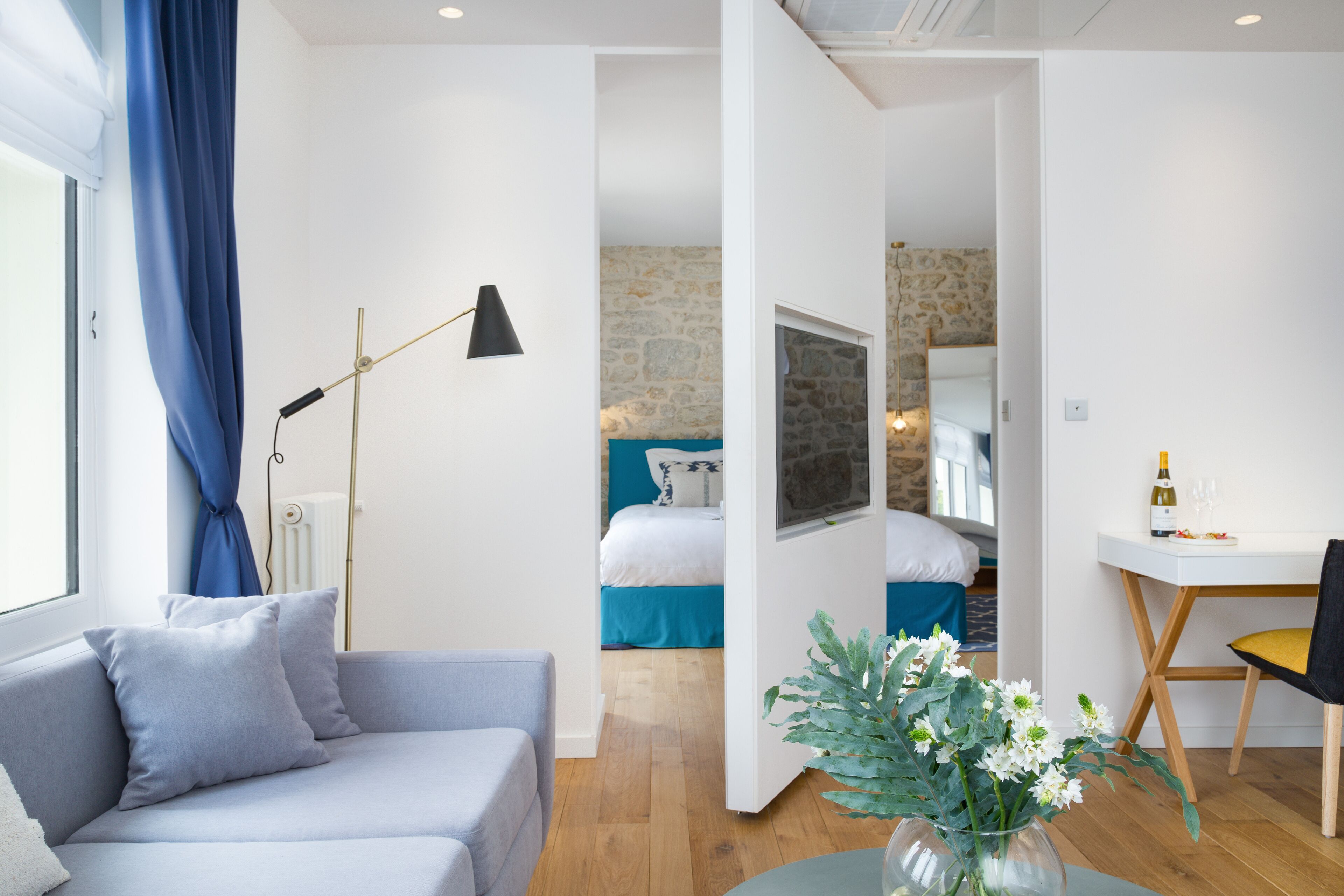 A modern hotel room with a grey sofa, a dining table with a chair, a vase with flowers, a bed with blue bedding, and a stone wall.