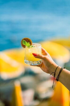 Una mano sostiene un vaso con una bebida adornada con una rodaja de fruta y una hoja de menta, sobre un fondo borroso con toques de azul y amarillo.
