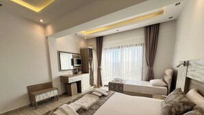 A hotel room with a large bed, a desk with a computer, a chair, a window with curtains, and a headboard with a mirror. - Sardunya Andifli Otel (Kas)