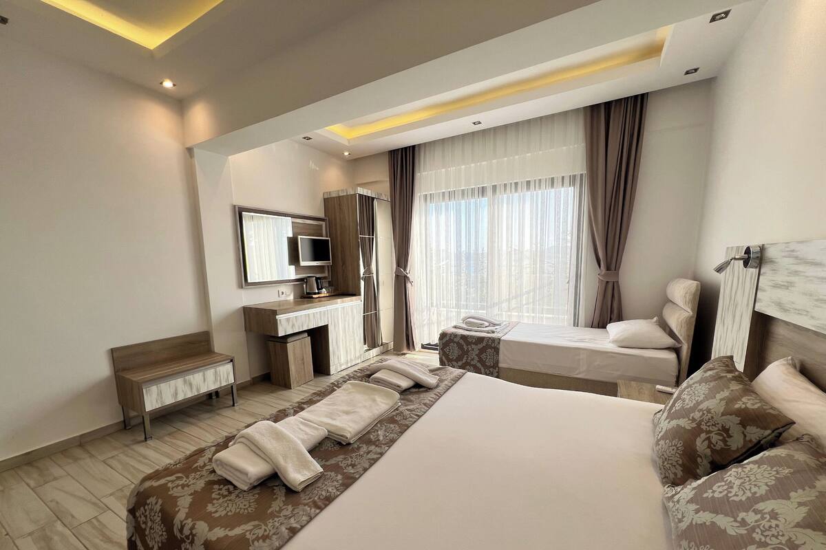 A hotel room with a large bed, a desk with a computer, a chair, a window with curtains, and a headboard with a mirror.