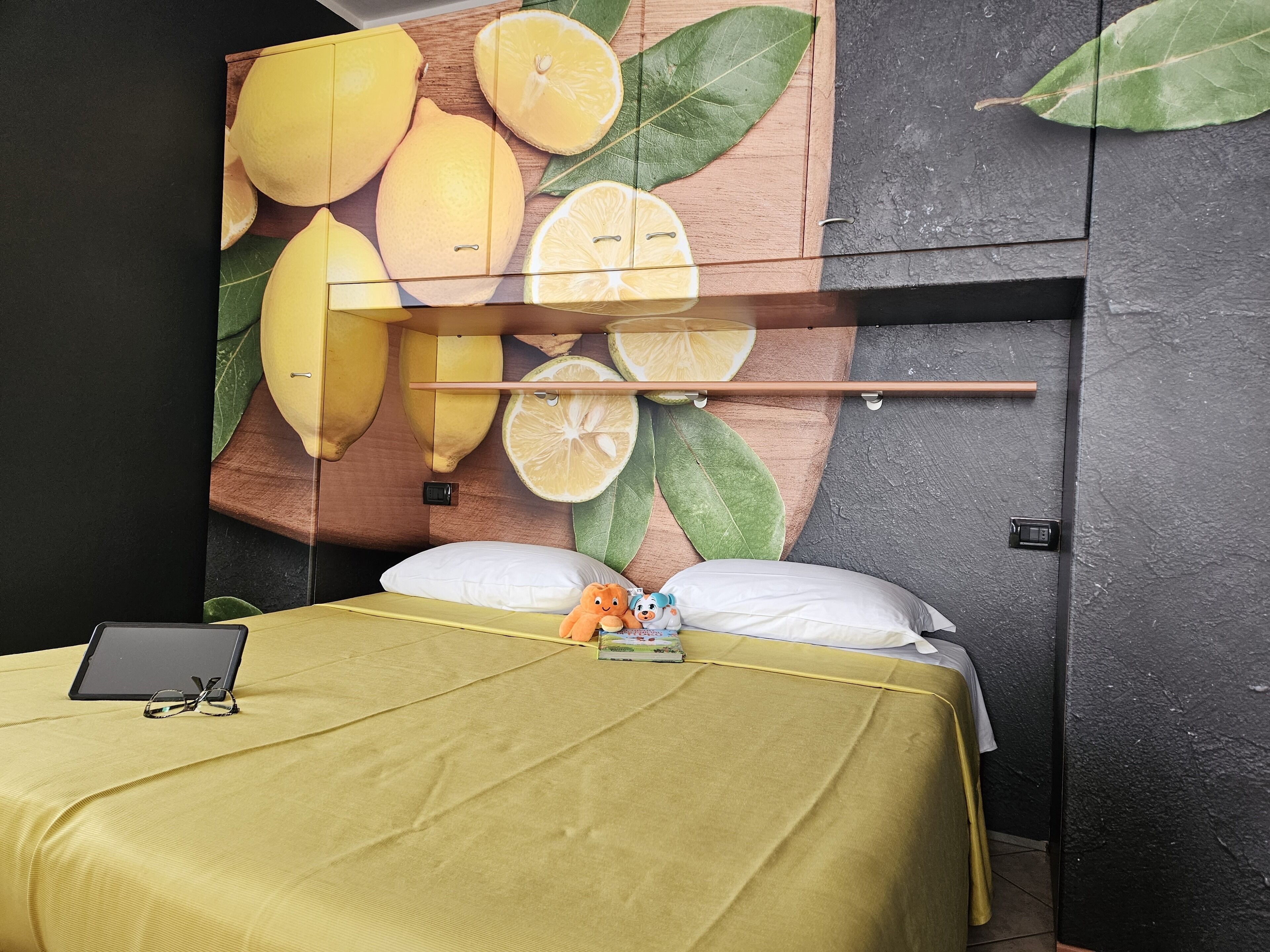 A bed with a yellow bedspread, two pillows, a tablet, and a stuffed toy on the bed.