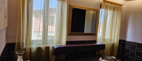 A room with a bed, a mirror, a window with curtains, a table with a glass of water and two apples, and a tiled wall.