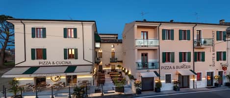 Un edificio con letreros que dicen 'PIZZA & CUCINA' y 'PIZZA & BURGER', mesas al aire libre y un balcón.