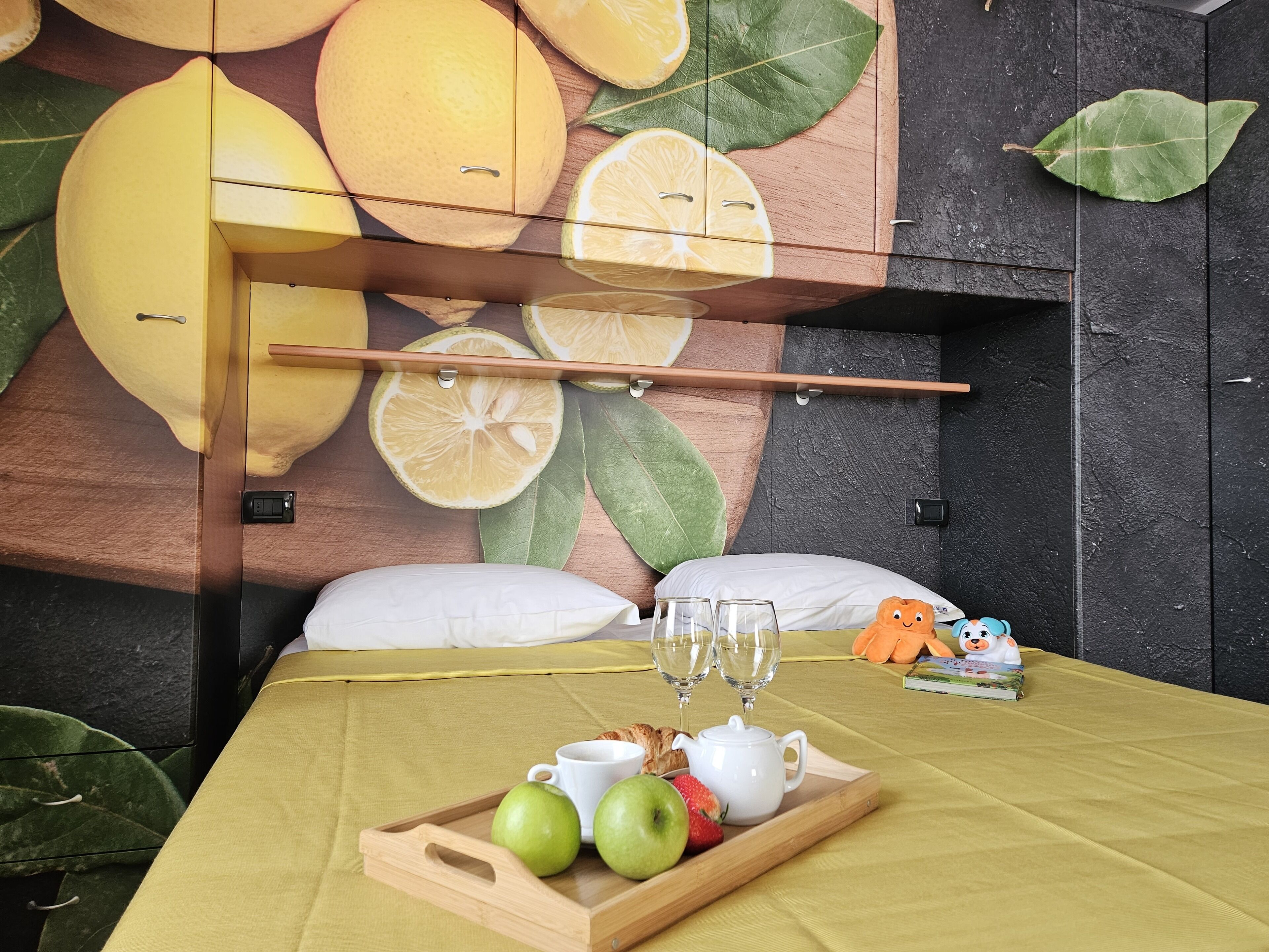 A hotel room with a bed, a breakfast tray with fruits and a teapot, and a wall mural of citrus fruits.