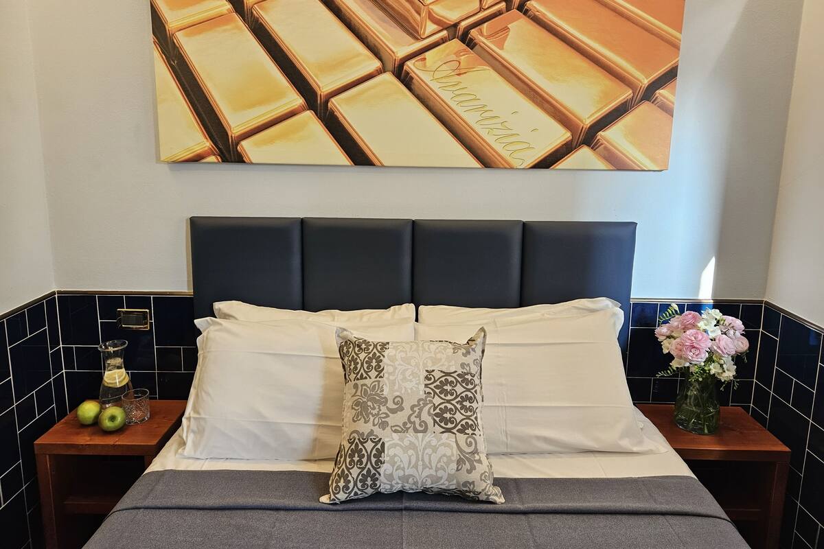 A neatly made bed with a dark headboard, white linens, and a patterned pillow. There is a small wooden nightstand with a glass and a bottle on it, and a vase with flowers on the right side.