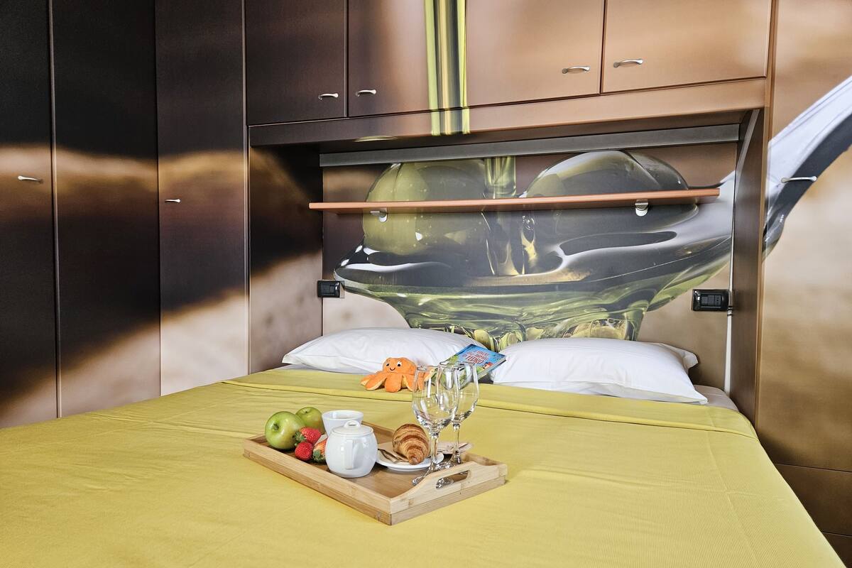 A hotel room with a bed, a tray with fruits, a croissant, a teapot, and a magazine.