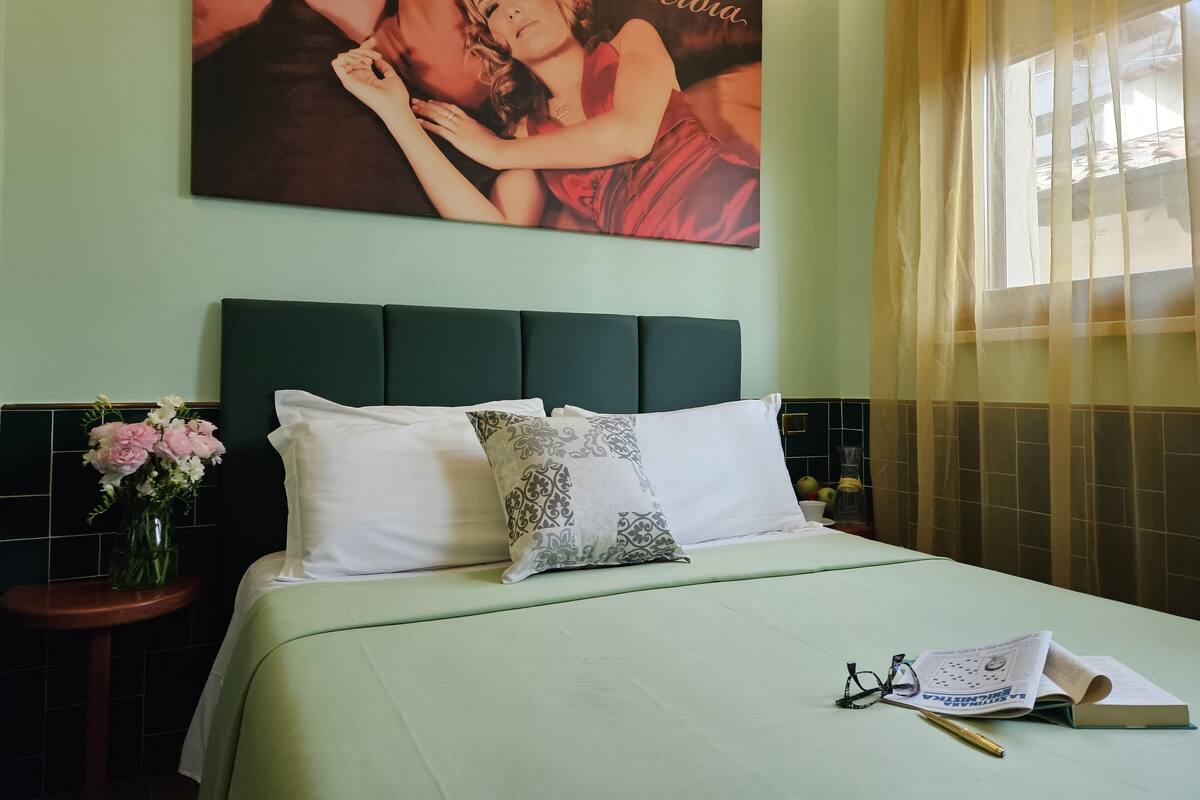 A neatly made bed with a green bedspread, white pillows, and a decorative pillow with a floral pattern. A vase with pink and white flowers is on a nightstand beside the bed. A large framed picture hangs on the wall above the bed.