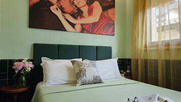 A neatly made bed with a green bedspread, white pillows, and a decorative pillow with a floral pattern. A vase with pink and white flowers is on a nightstand beside the bed. A large framed picture hangs on the wall above the bed.