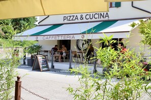 Un café avec une terrasse, une enseigne indiquant « PIZZA & CUCINA », et un panneau affichant le menu.