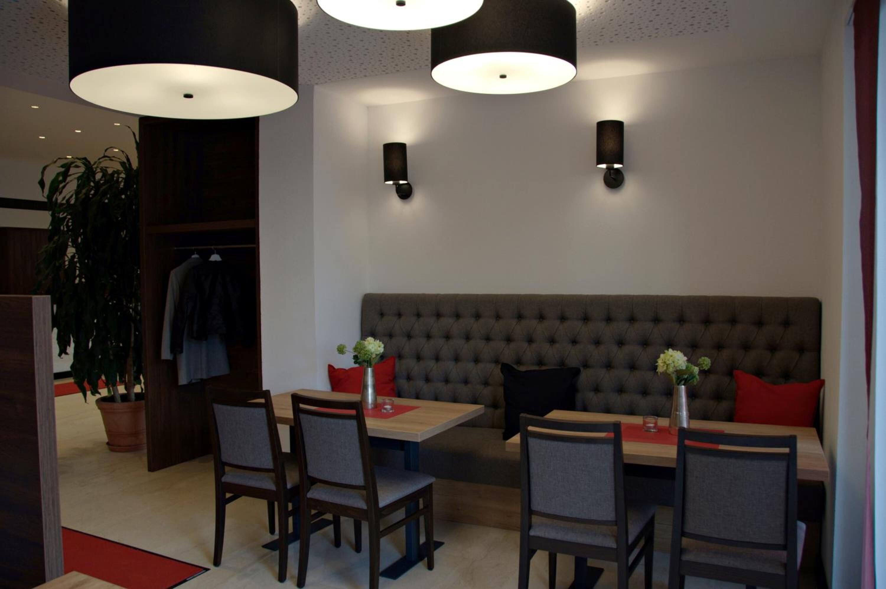A modern dining area with a sofa, chairs, and a table. There are pendant lights above the table and wall-mounted lights. A potted plant is visible in the corner.