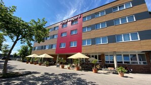 A multi-story hotel with a red and brown facade, outdoor seating area with umbrellas, and potted plants. - Hotel Asgard (Gersthofen)
