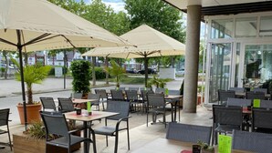An outdoor cafe with tables and chairs, umbrellas, and potted plants. - Hotel Asgard (Gersthofen)