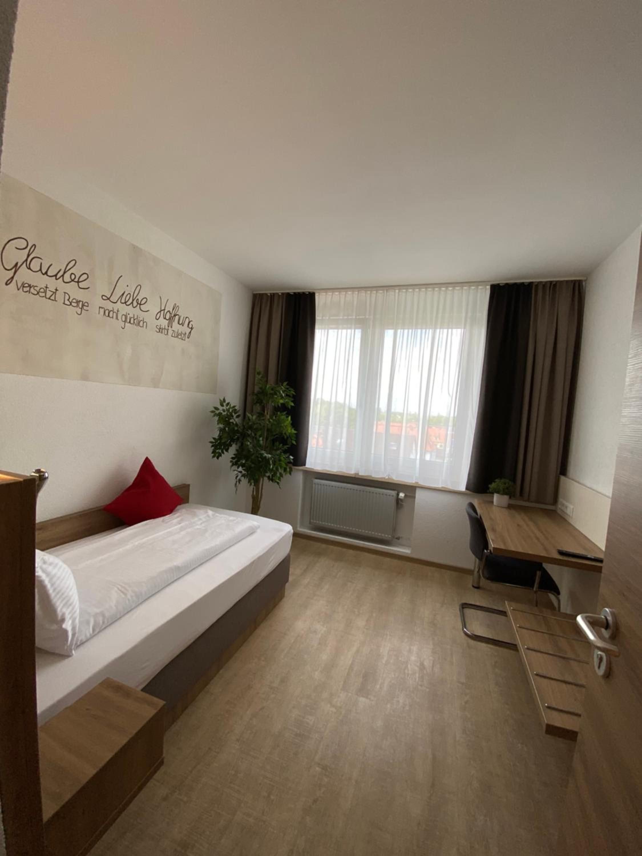 Habitación de hotel con cama, escritorio, silla, planta en maceta y ventana con cortinas.