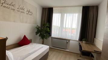 Habitación de hotel con cama, escritorio, silla, planta en maceta y ventana con cortinas.