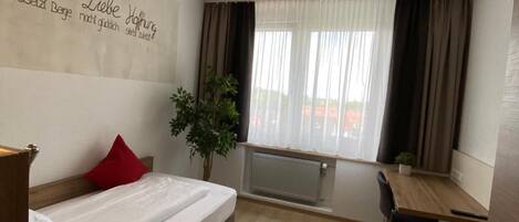 Habitación de hotel con cama, escritorio, silla, planta en maceta y ventana con cortinas.