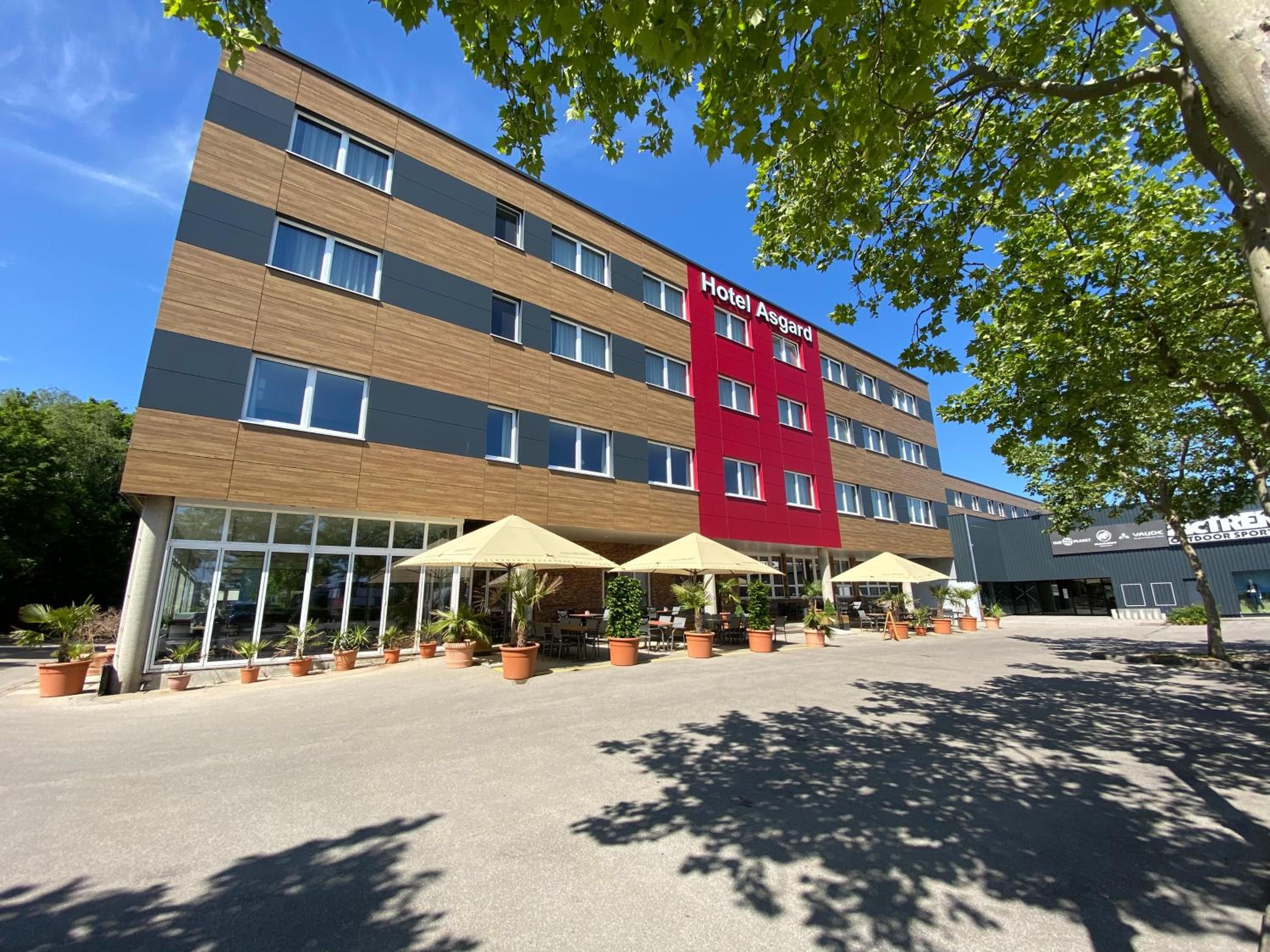 Un hotel con fachada roja y marrón, múltiples ventanas y zona de estar exterior con sombrillas.