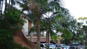 Un edificio de ladrillo con una escalera que lleva a un balcón. Se observan vehículos estacionados y abundante vegetación alrededor.