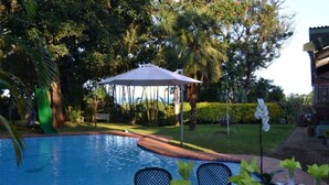 Zona junto a la piscina con mesa y sillas, rodeada de exuberante vegetación y una piscina azul cristalina.